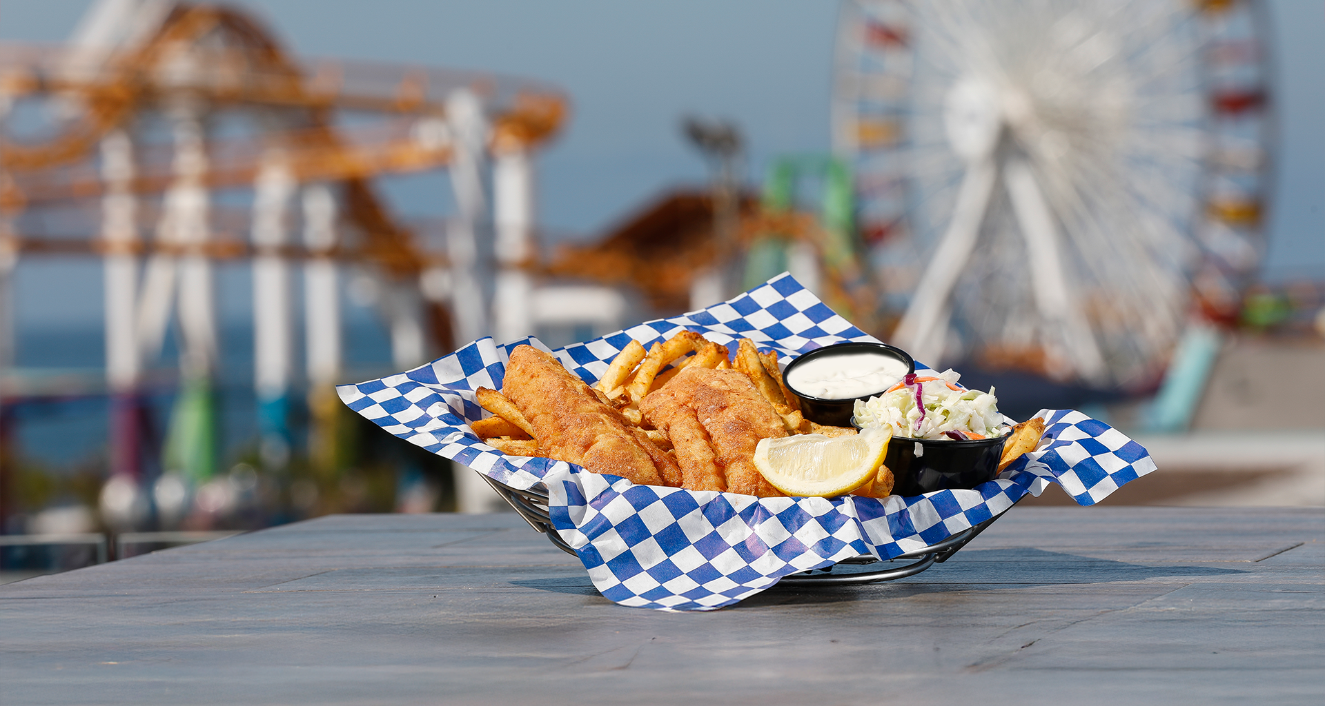 Golden-brown fish and chips served with coleslaw, tartar sauce, and lemon at Seaside on the Pier, with a Ferris wheel in the background.