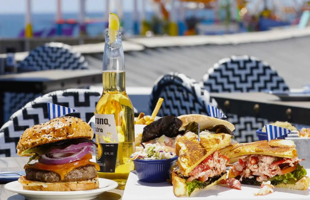 Burger with cheddar, lettuce, tomato, and red onion alongside a lobster grilled cheese sandwich and fries, served with a bottled beer on an outdoor patio at Seaside on the Pier.